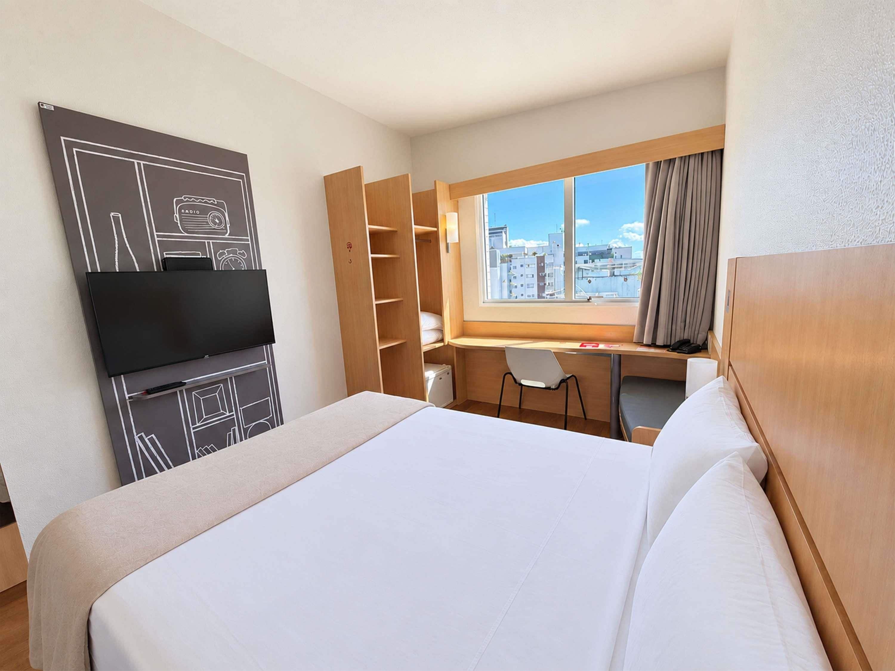 Habitación de hotel con cama, escritorio, silla, televisor y un ventanal con vista a la ciudad.