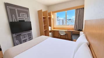 Habitación de hotel con cama, escritorio, silla, televisor y un ventanal con vista a la ciudad.