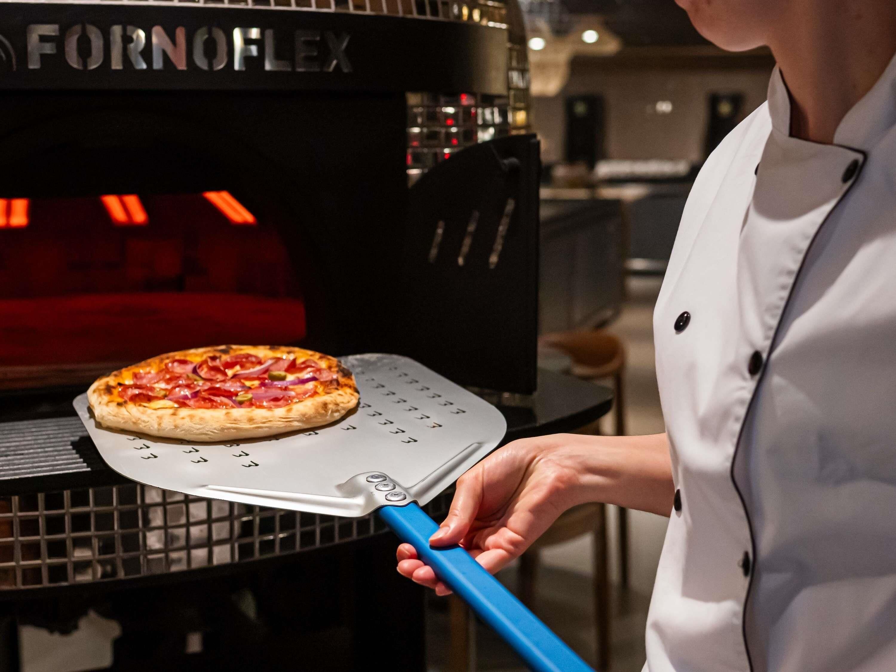 A person holding a pizza peel in front of a commercial pizza oven.