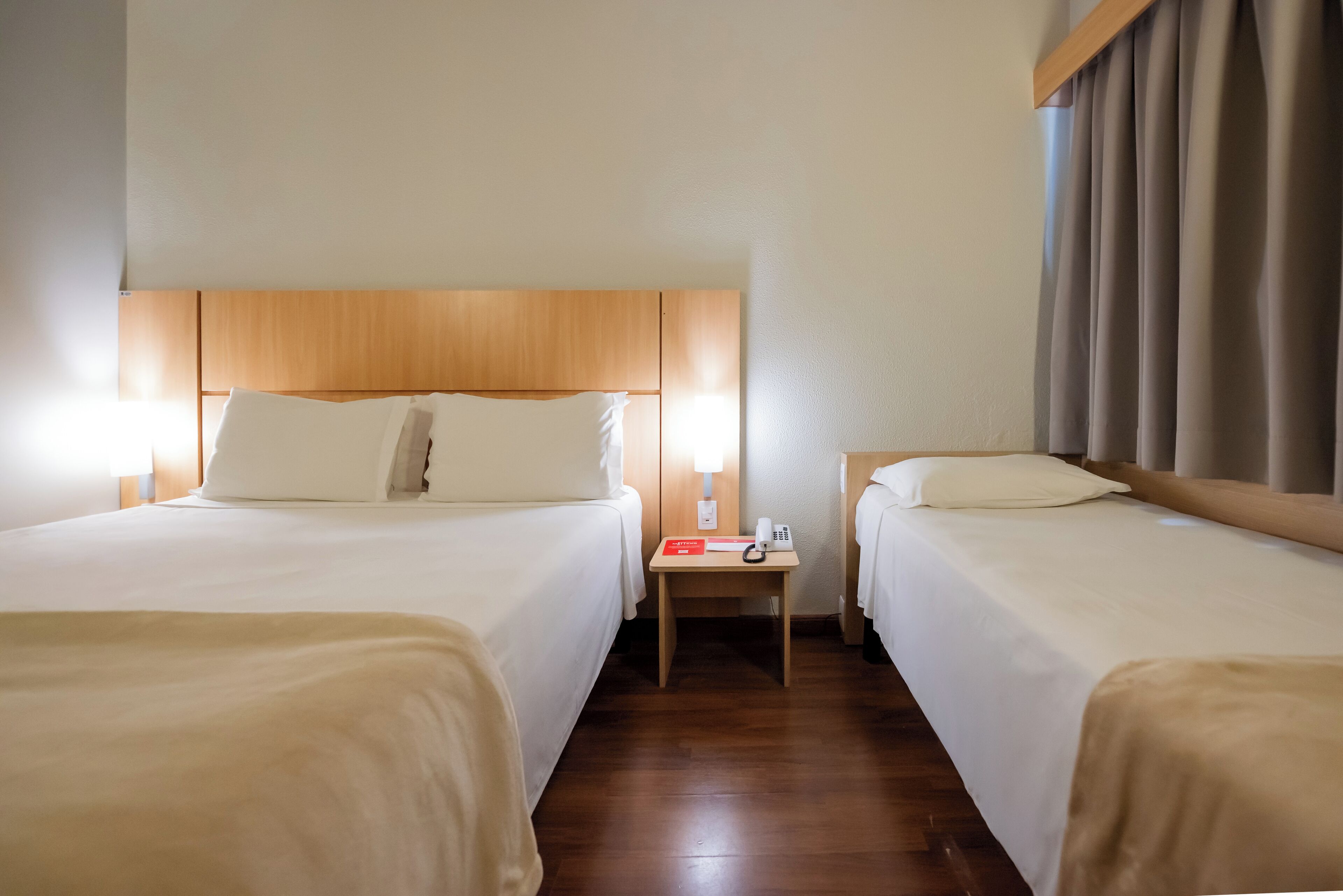 A hotel room with two beds, a wooden headboard, a nightstand with a lamp, and a window with curtains.