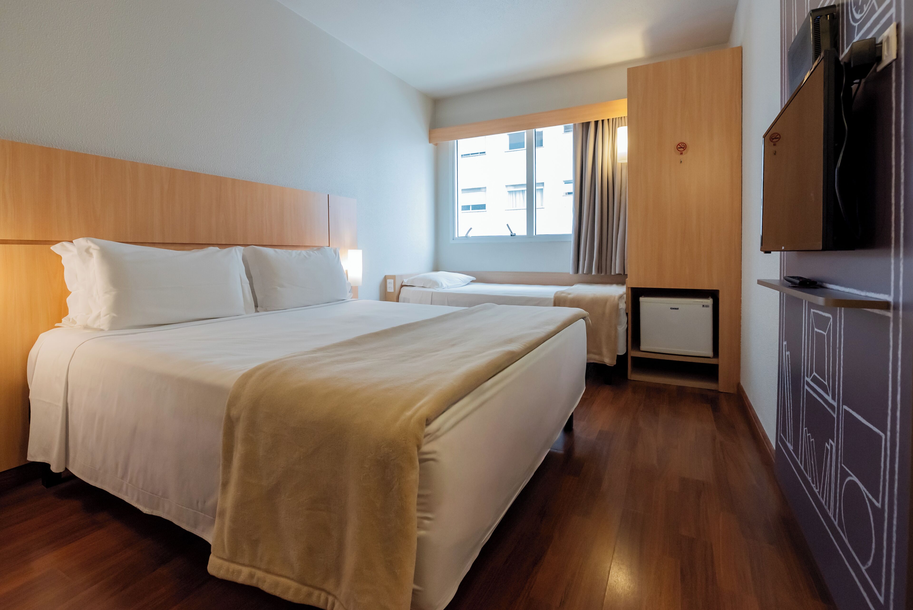 Habitación de hotel con una cama grande, una cama más pequeña, un armario, un televisor y una ventana con cortinas.