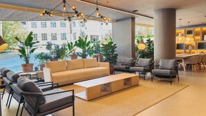 A modern lounge area with a sofa, chairs, and a coffee table, featuring large windows with a view of buildings and plants outside.