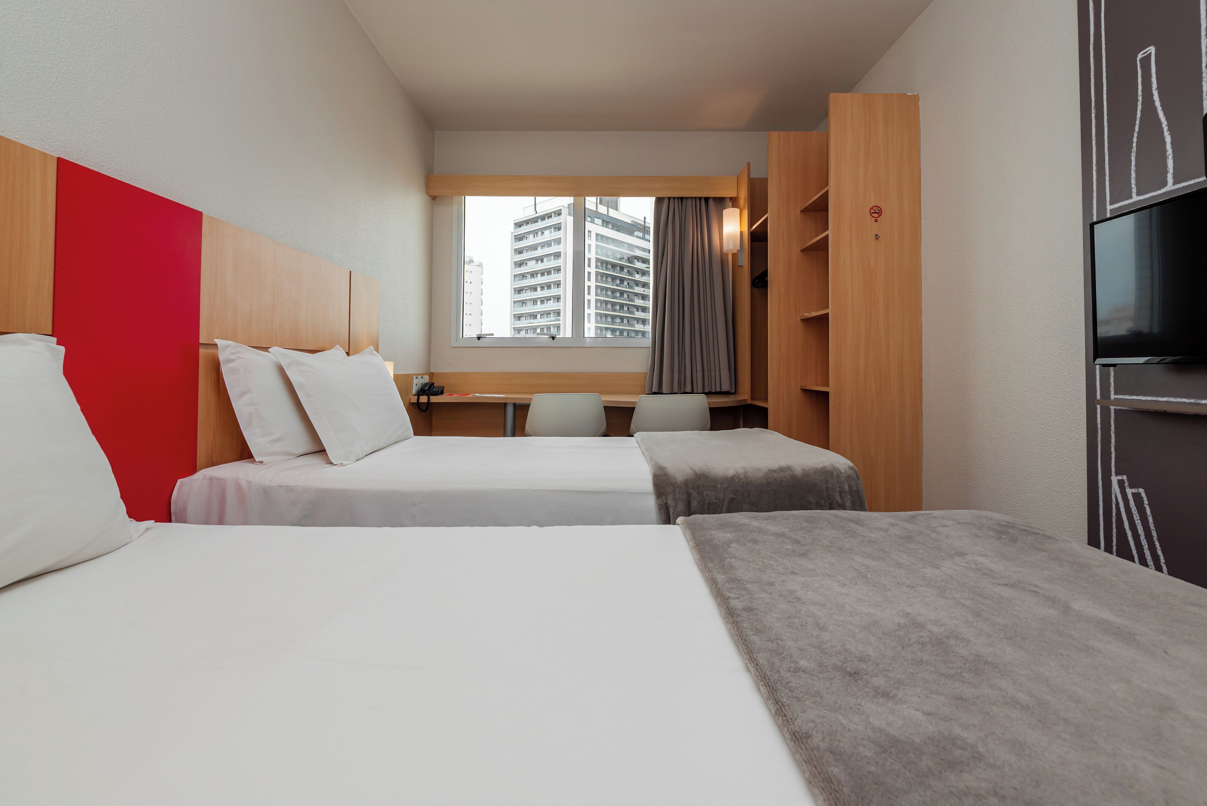 A hotel room with two beds, a TV, a desk, and a window with a city view.