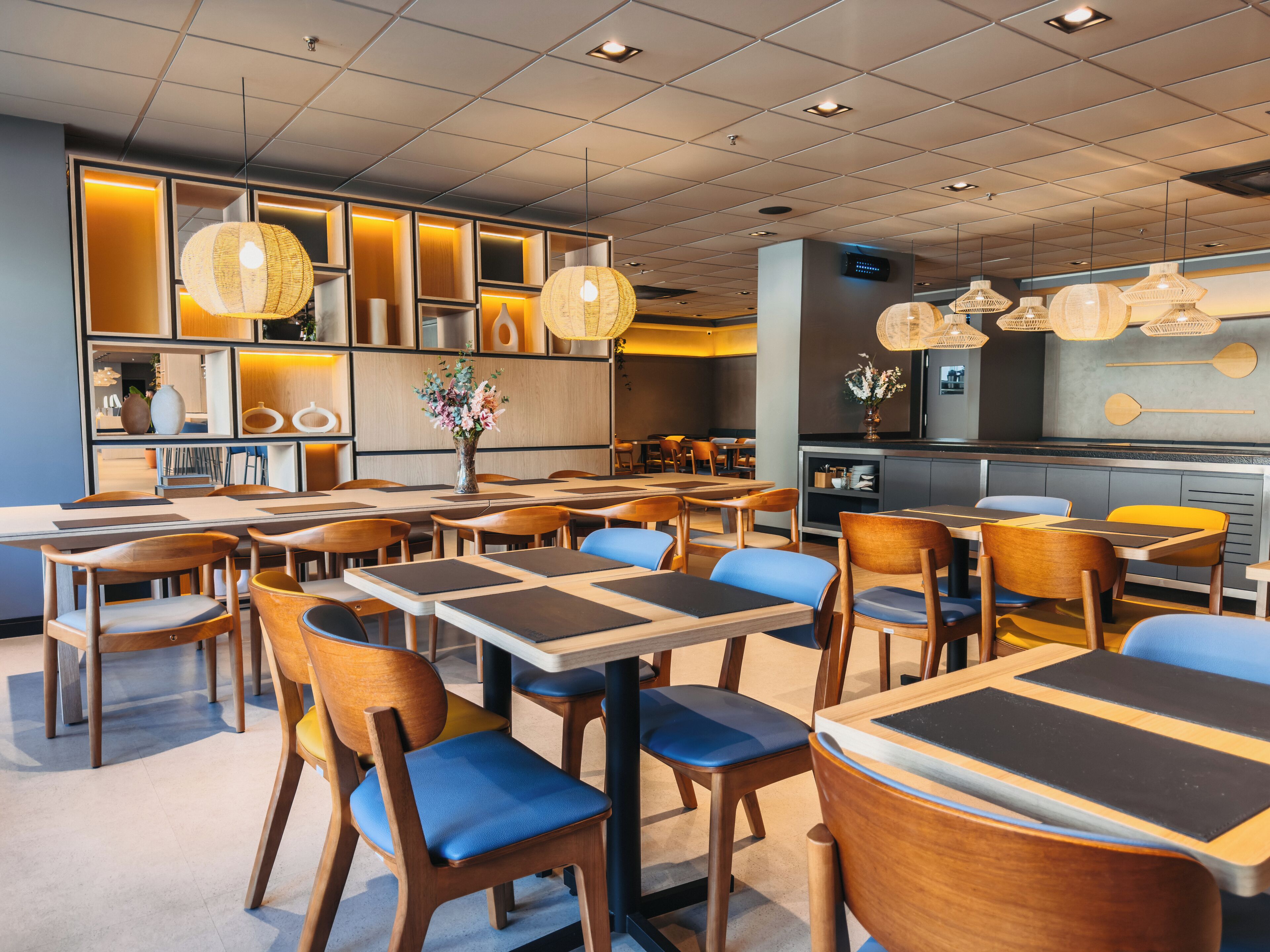 A modern restaurant with wooden tables and chairs, a central floral arrangement, and hanging wicker lamps.