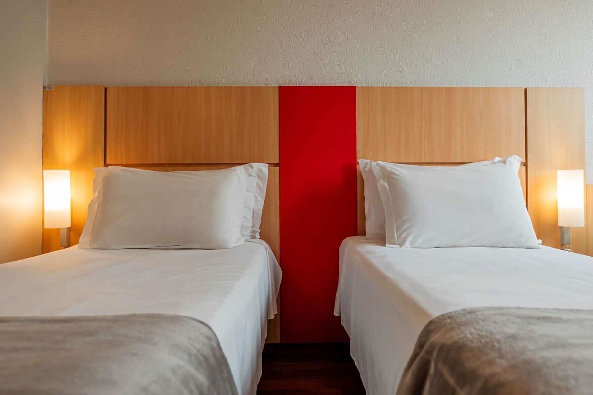 Una camera d'albergo con due letti, testiere in legno, una parete di colore rosso e lampade sui comodini.