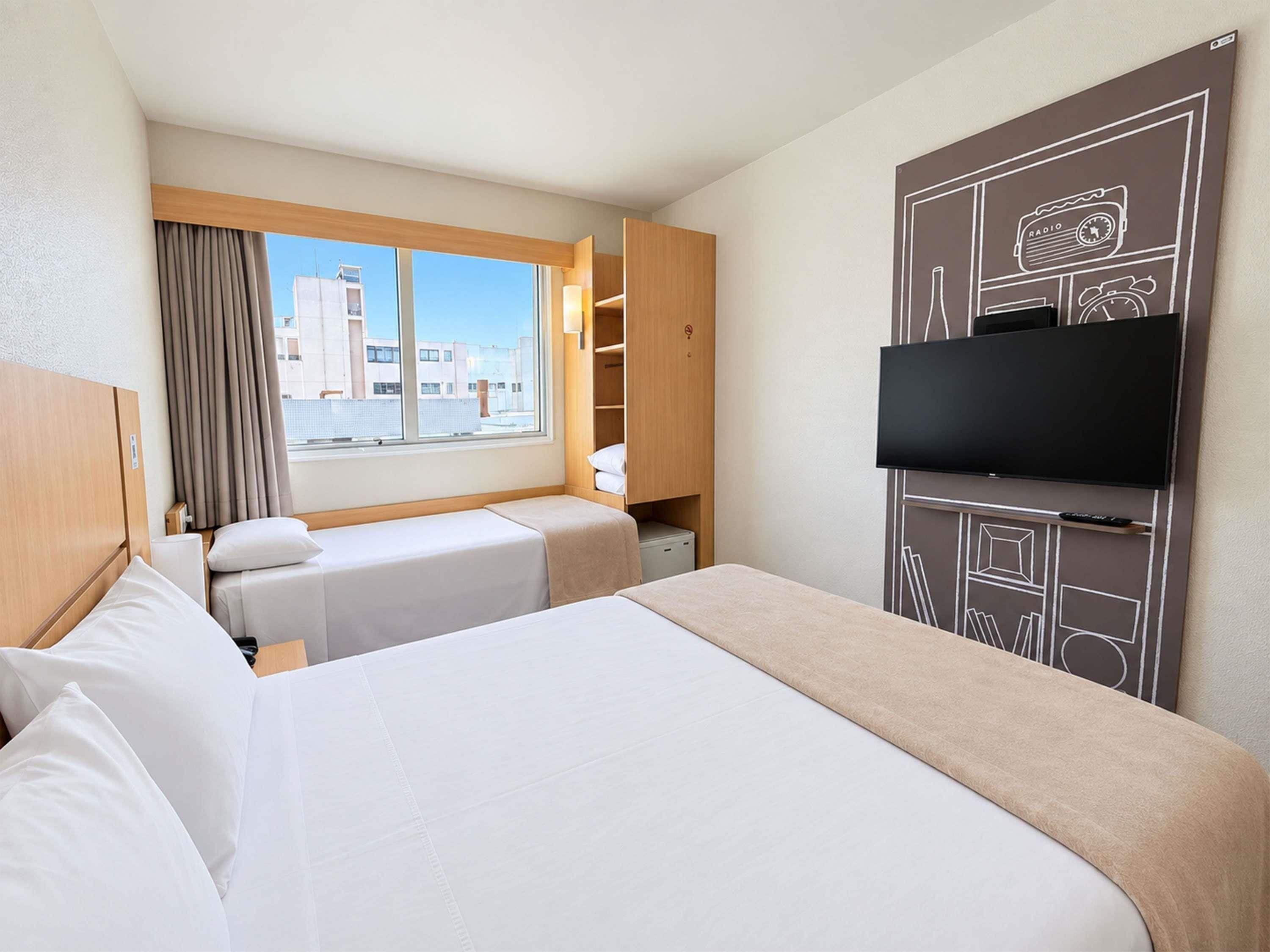 A hotel room with two beds, a TV, a window with curtains, and a view of buildings.