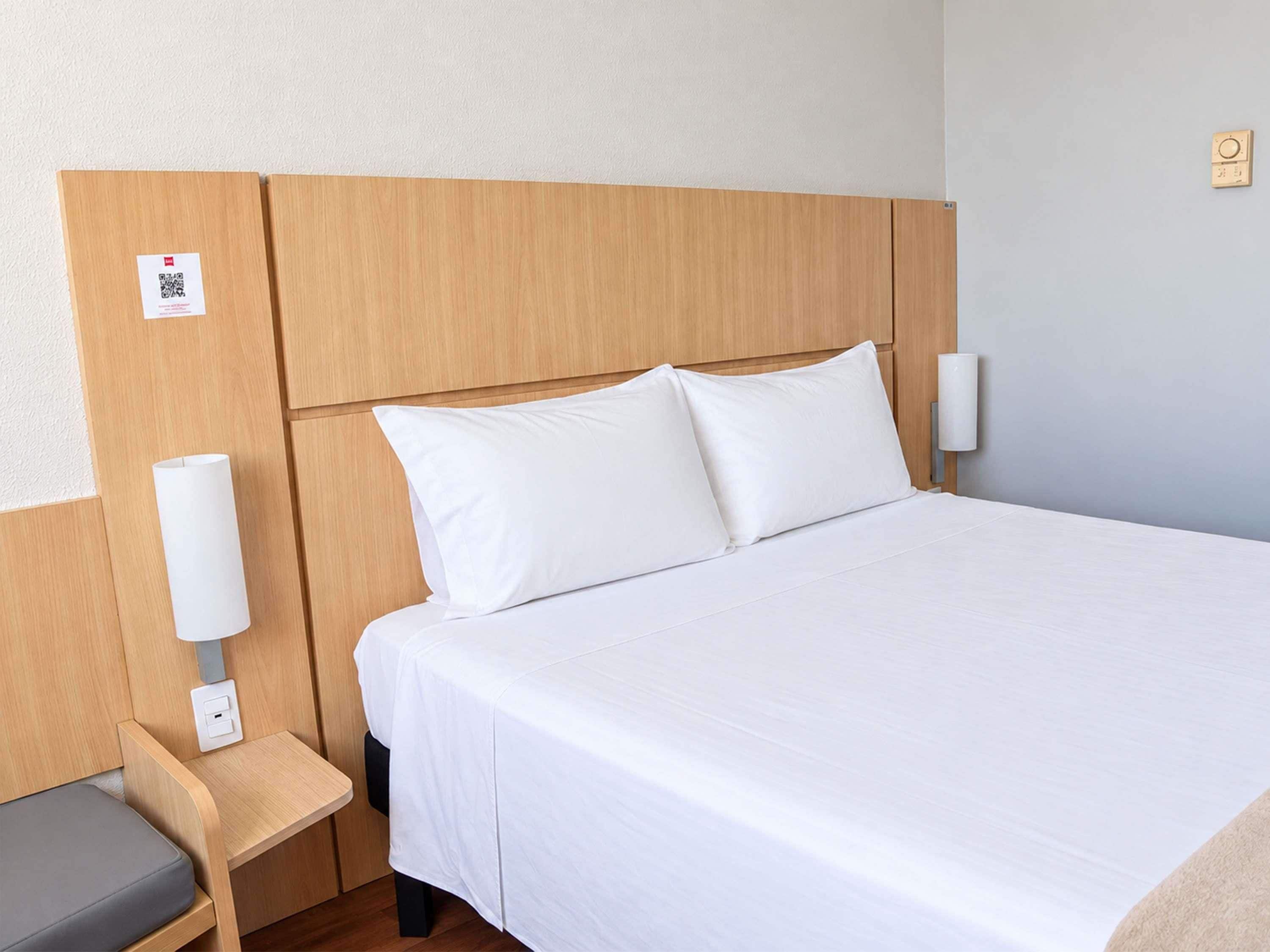 A hotel room with a large bed, a bedside table, a chair, and a wall-mounted light fixture.