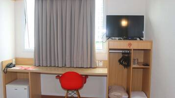 A room with a desk, a television, a red chair, a small refrigerator, and a wardrobe with hanging items.