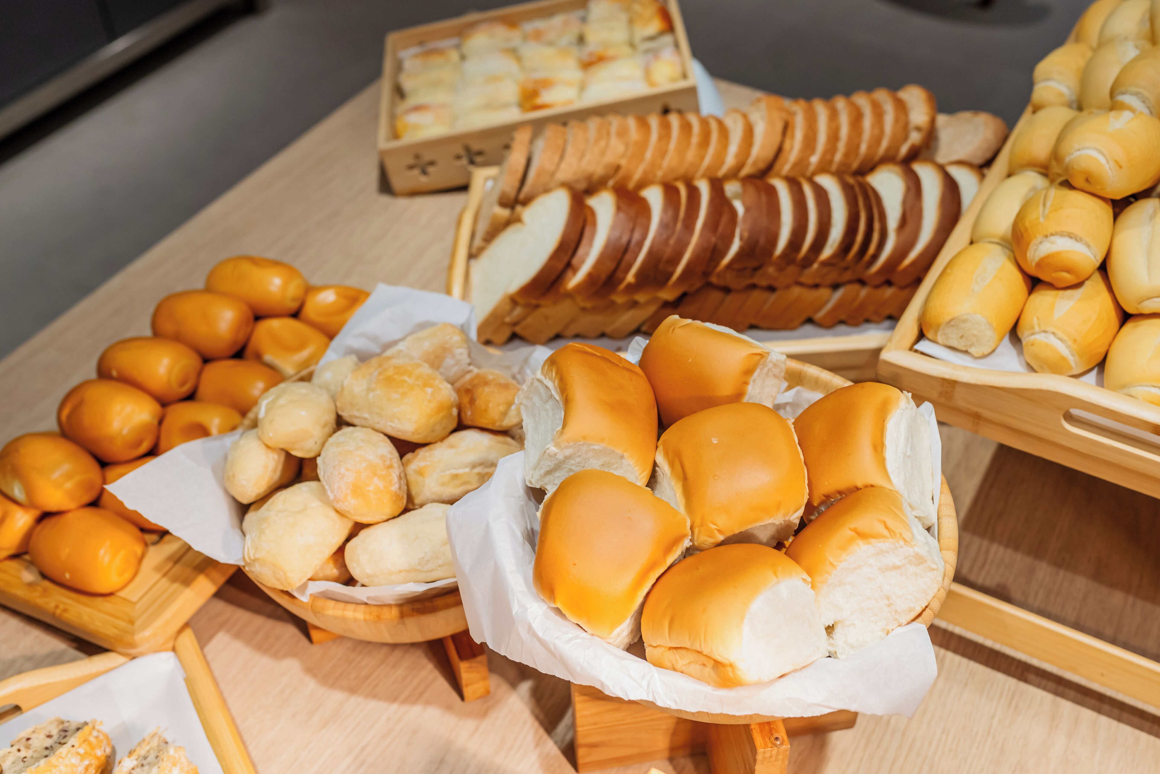 Diversos produtos de panificação, como pão, pães de forma e doces, expostos em suportes de madeira.