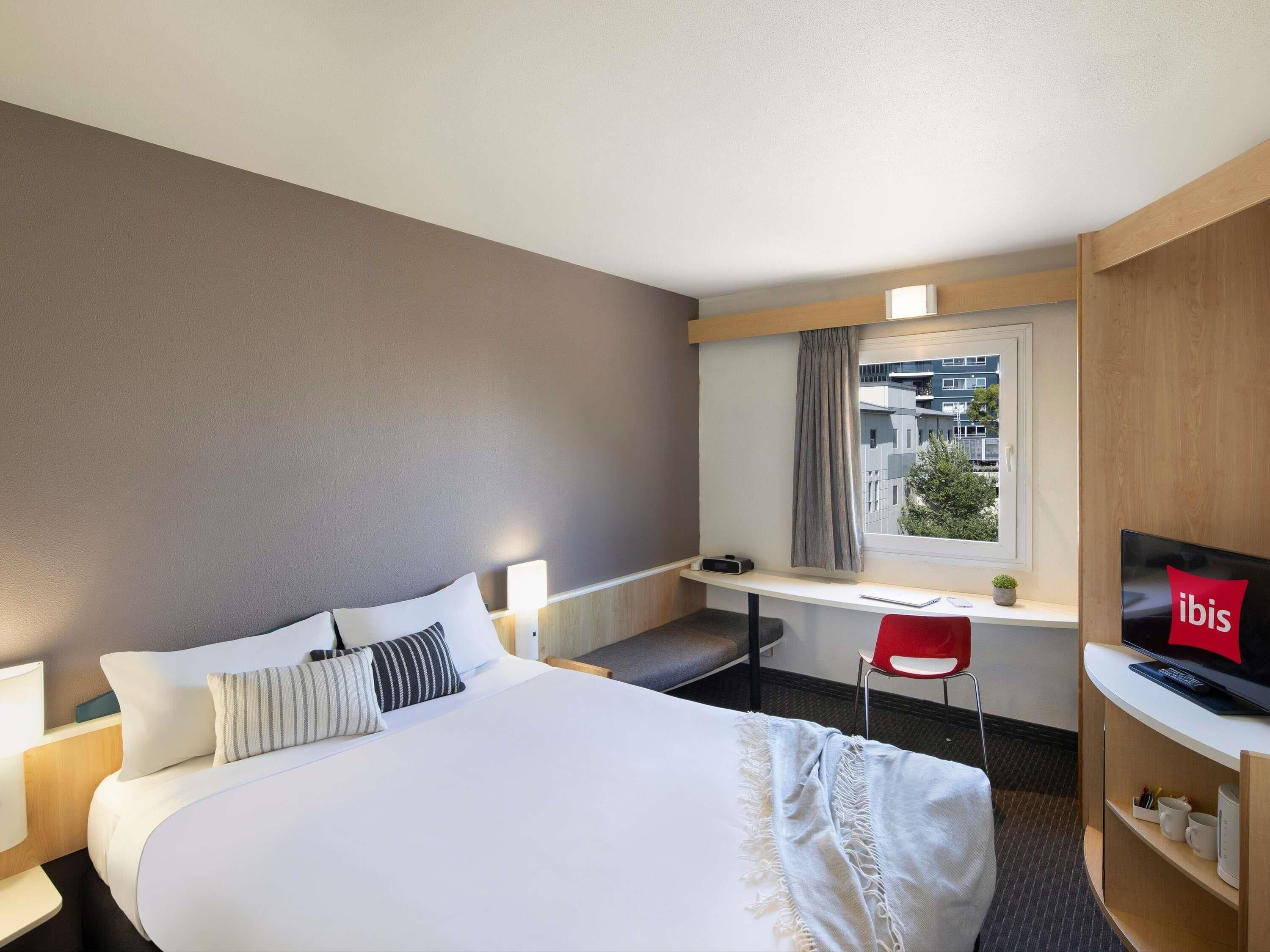 A hotel room with a bed, a desk, a chair, a TV, and a window with a view of buildings.