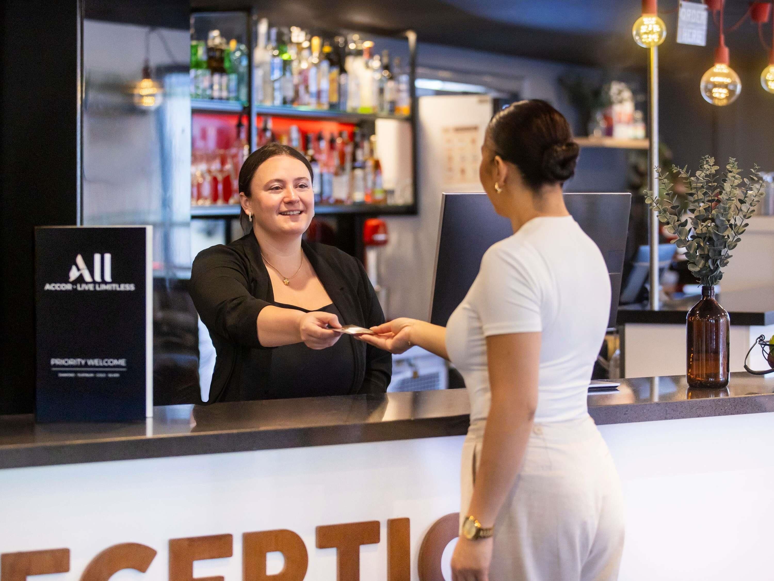 Una zona de bar con mostrador, un cartel que dice 'All Priority Welcome' y una estantería con diferentes botellas.