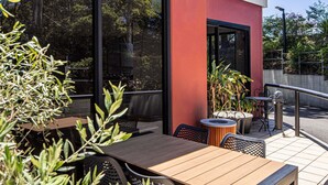 Un patio al aire libre con mesas y sillas de madera, rodeado de follaje y una pared roja.