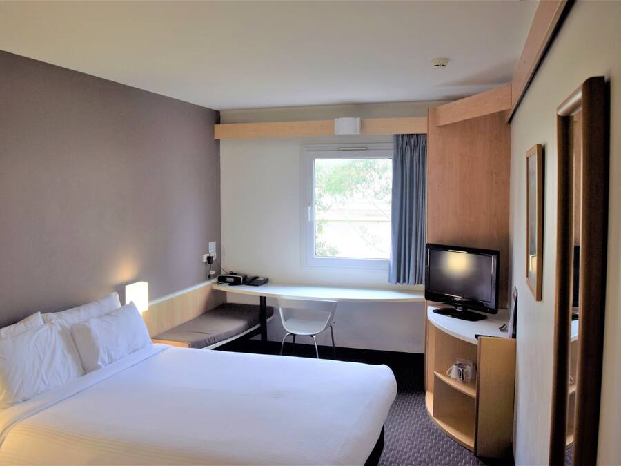 Una habitació d'hotel amb un llit, una taula de treball amb una cadira, una televisió i una finestra amb cortines.