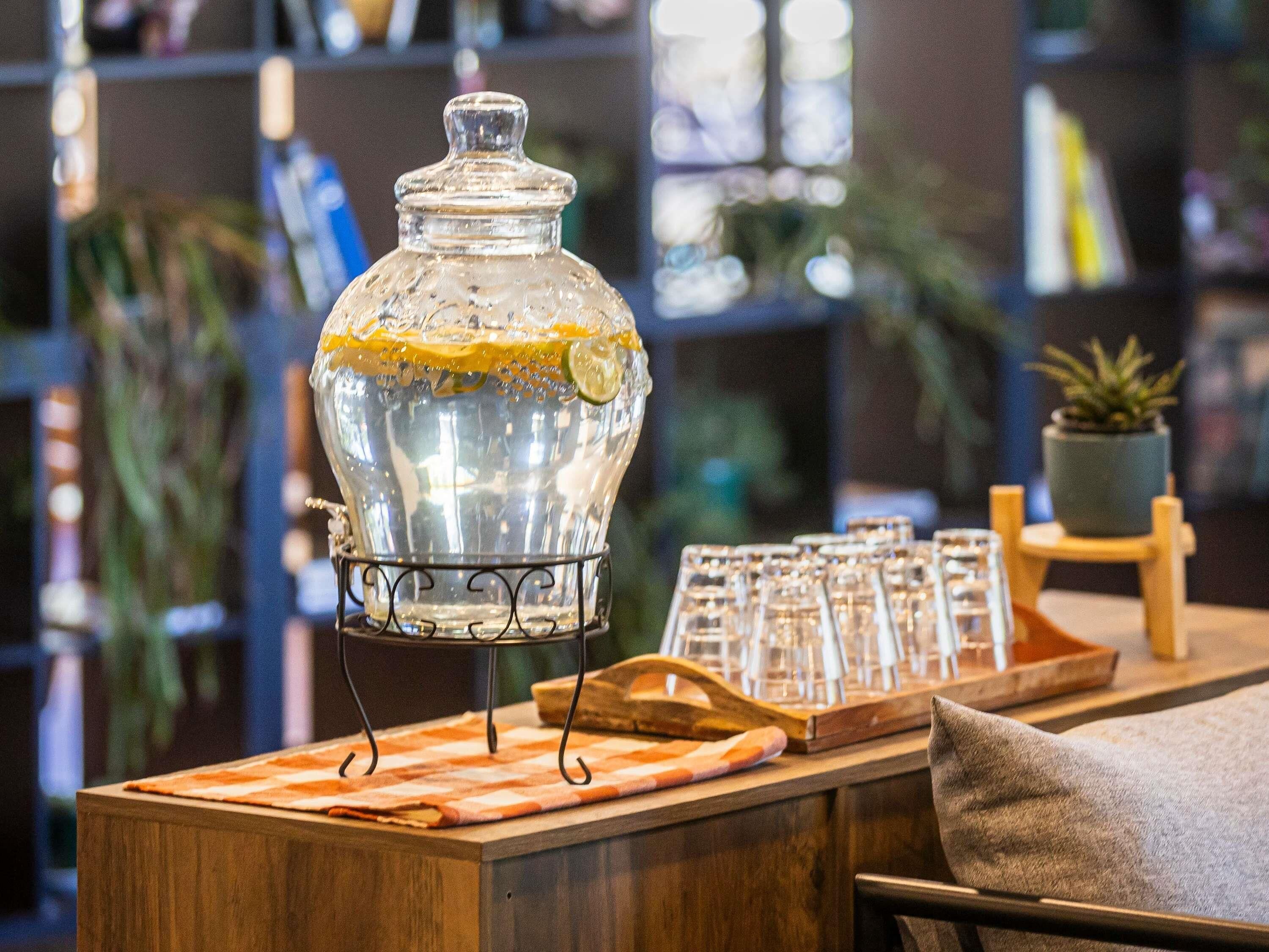 Une carafe en verre avec une tranche de citron posée sur une table en bois, entourée de verres et d’une petite plante en pot.