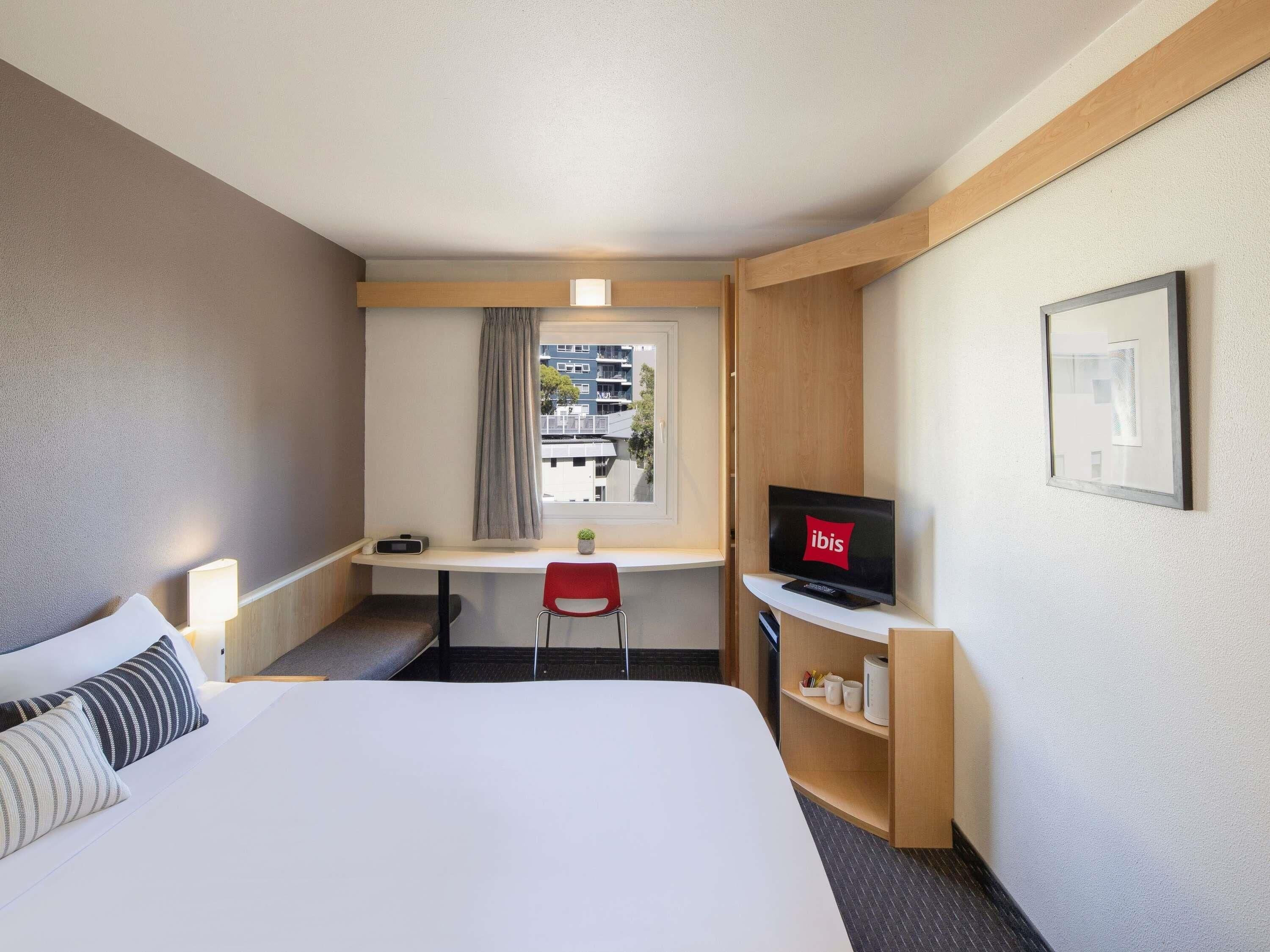 A hotel room with a bed, a desk with a computer, a TV, and a window with curtains.