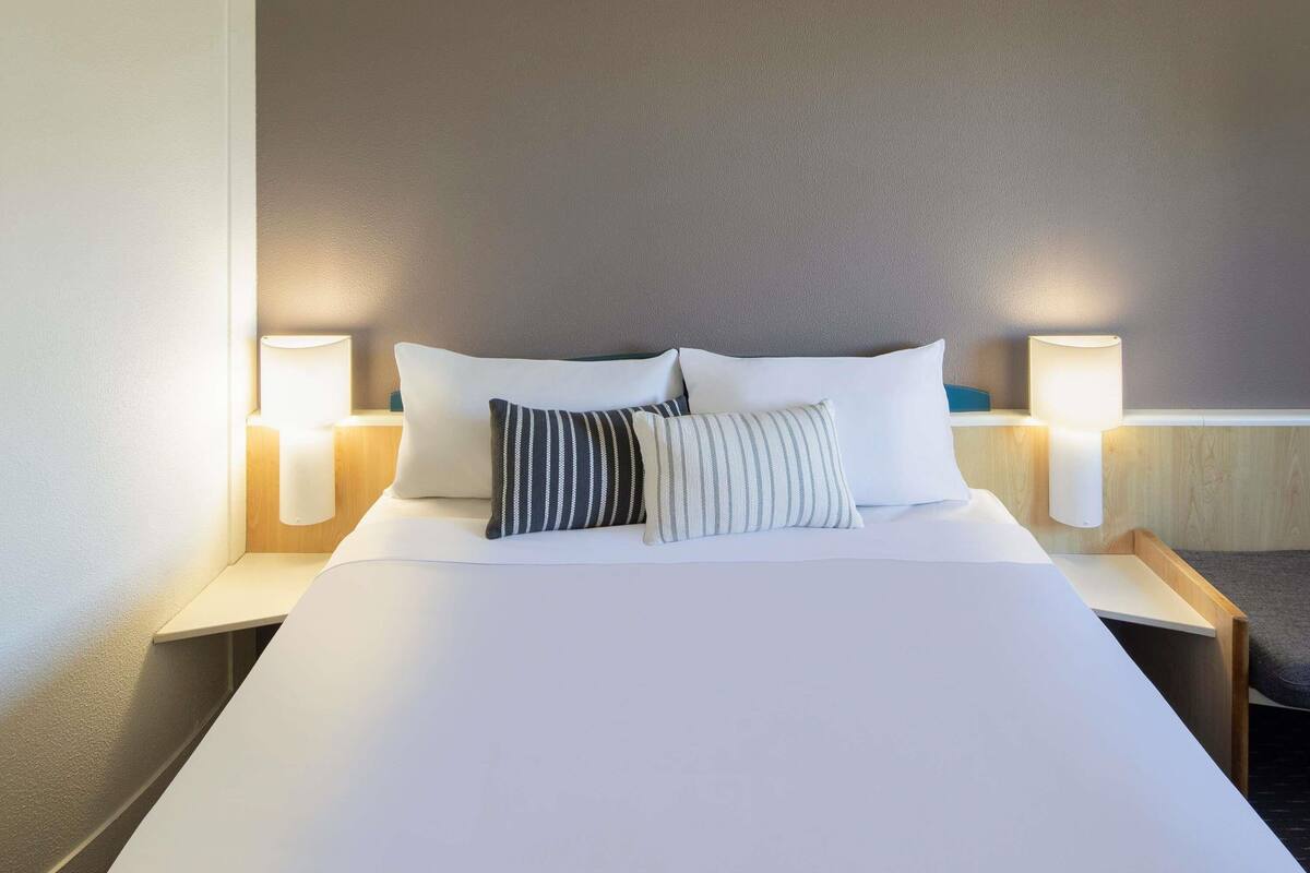 A neatly made bed with white linens and two pillows, flanked by two bedside lamps on wooden nightstands.