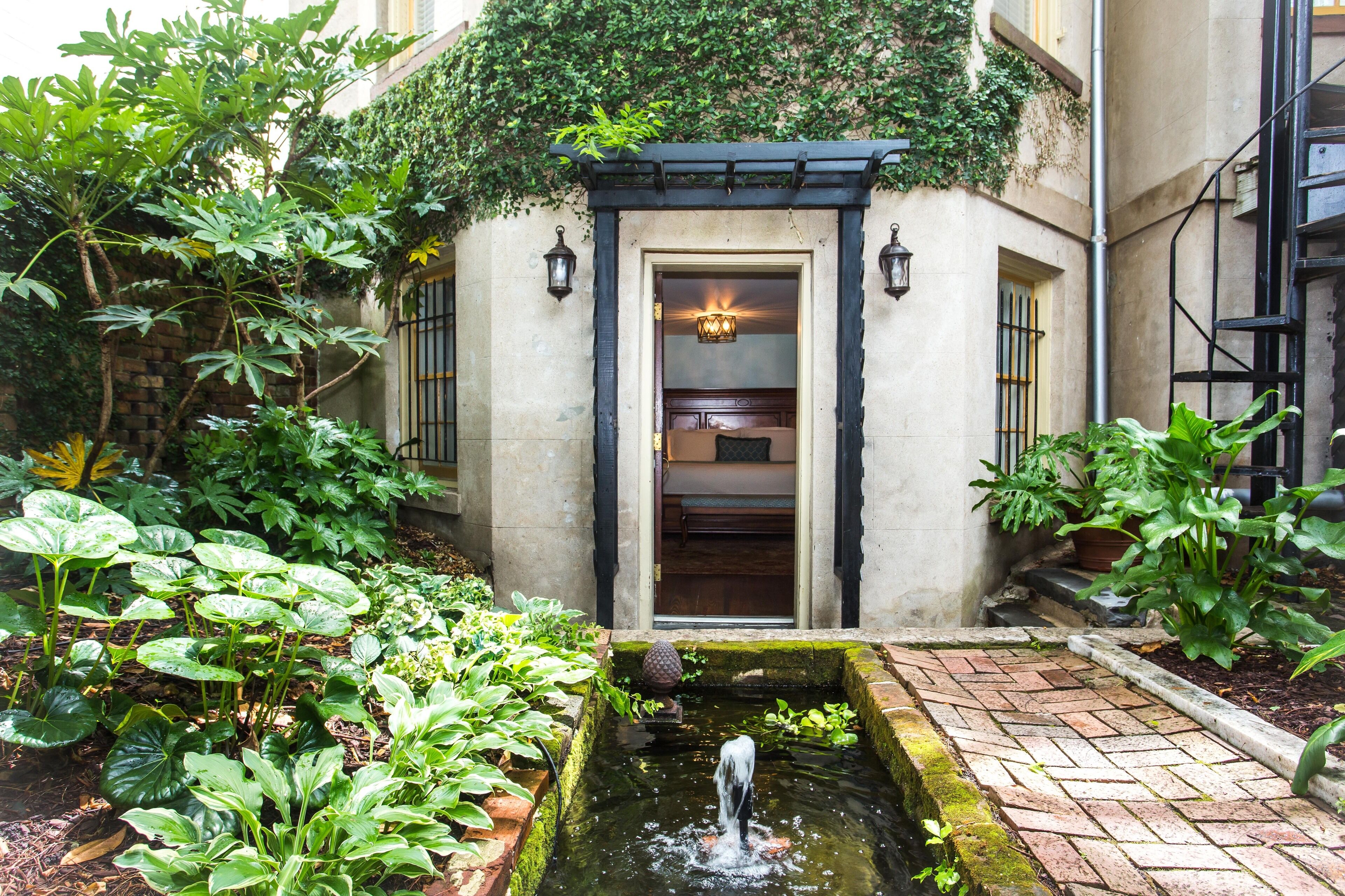 Une cour agrémentée d’un petit élément décoratif aquatique, d’une végétation luxuriante et d’une entrée de bâtiment avec une porte et deux lampes fixées au mur.