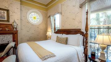 A neatly made bed with a white bedspread and a beige throw, a wooden headboard, a round mirror, and a window with a view of the outdoors.