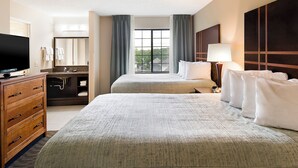 A hotel room with a large bed, a TV, a bathroom with a sink and mirror, and a window with curtains. - Aspen Suites (Rochester)