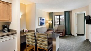 A hotel room with a kitchenette, a dining area with striped chairs, a sofa, a TV, and a window with curtains.