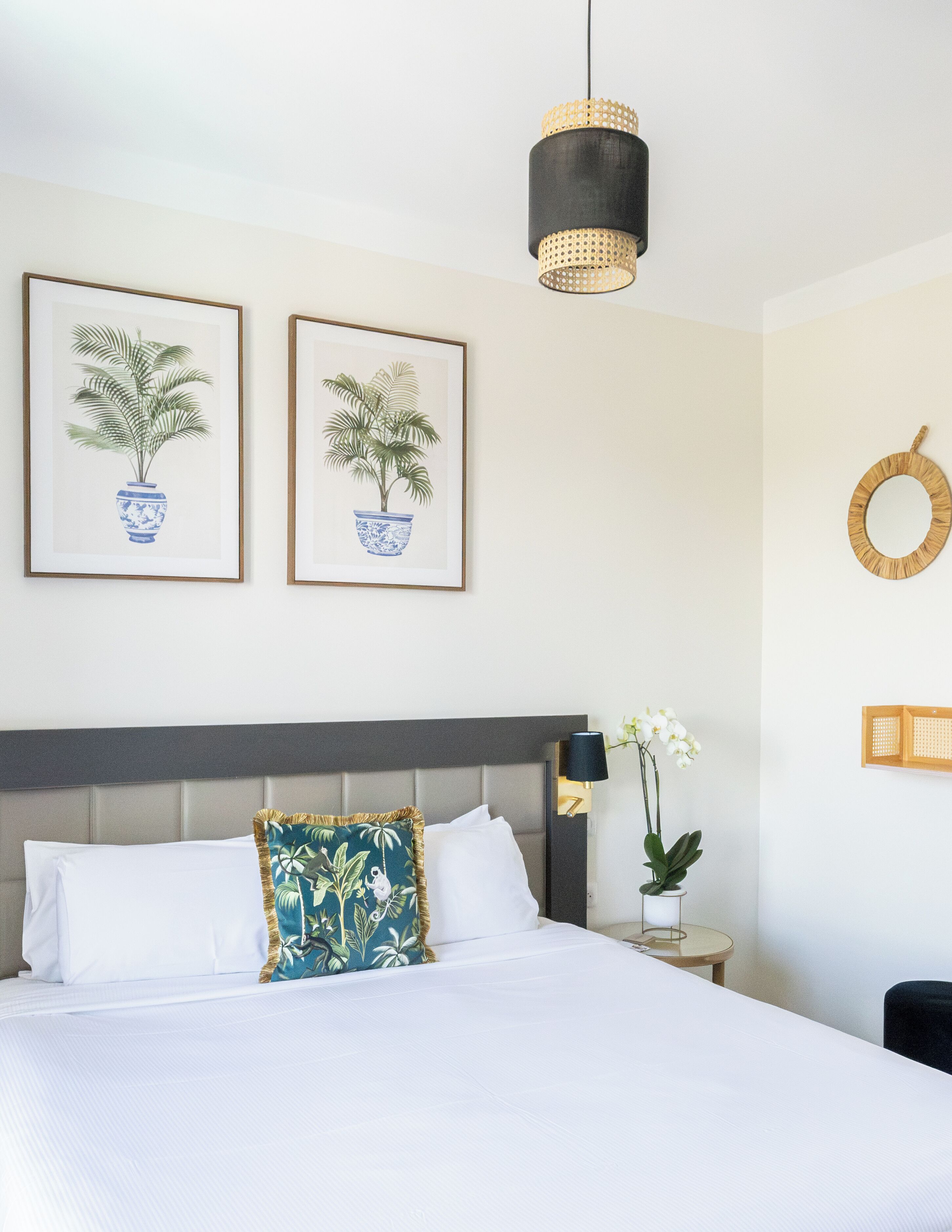 A neatly made bed with white linens, a grey headboard, and two framed botanical prints on the wall.