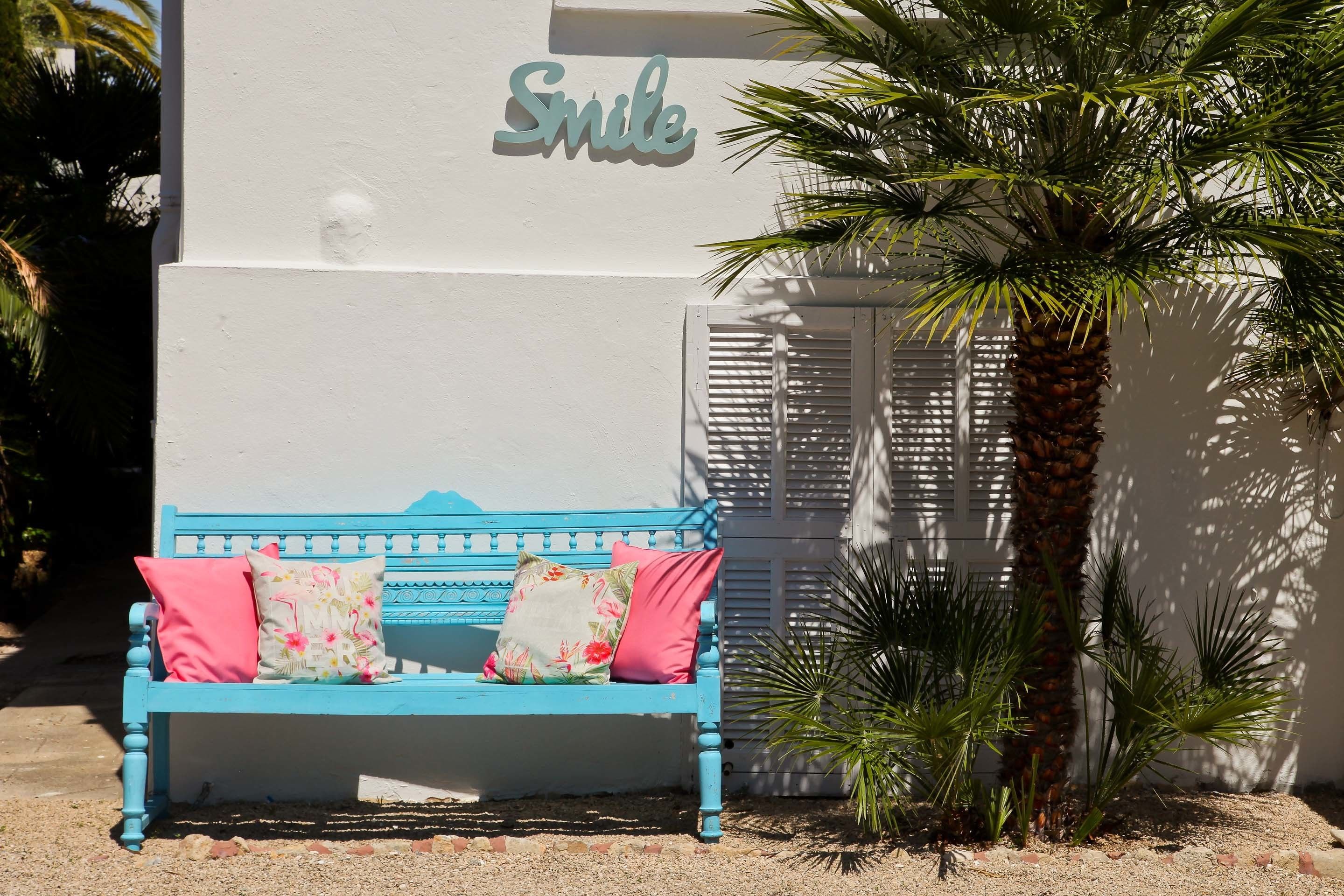 A blue bench with pink and floral cushions in front of a building with a 'Smile' sign.