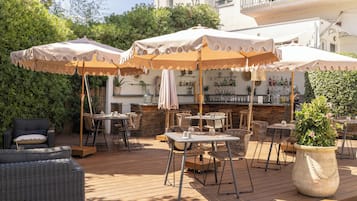 An outdoor patio area with multiple tables and chairs, some under umbrellas, and a bar area in the background.