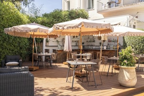 Un patio al aire libre con varias mesas y sillas, algunas bajo sombrillas, y un área de bar al fondo.
