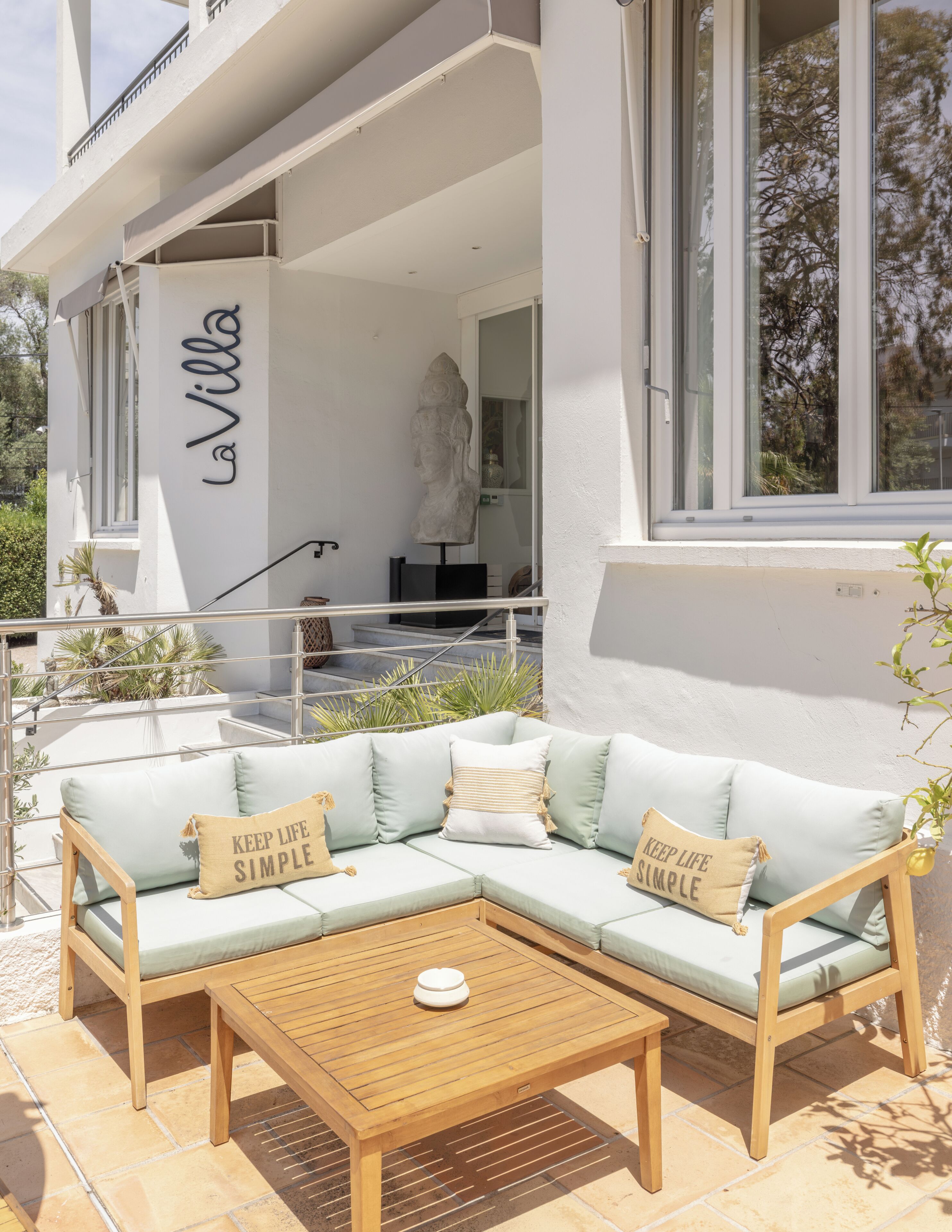 Un espacio exterior moderno con una zona de estar, un sofá de color claro y una mesa de madera, una escultura de piedra, y un edificio con un letrero que dice 'La Villa'.