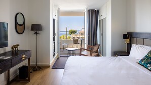 A hotel room with a bed, a desk, a chair, a TV, a lamp, and a balcony with a view of the sea.