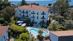 A multi-story building with a swimming pool, surrounded by greenery and a view of the sea.