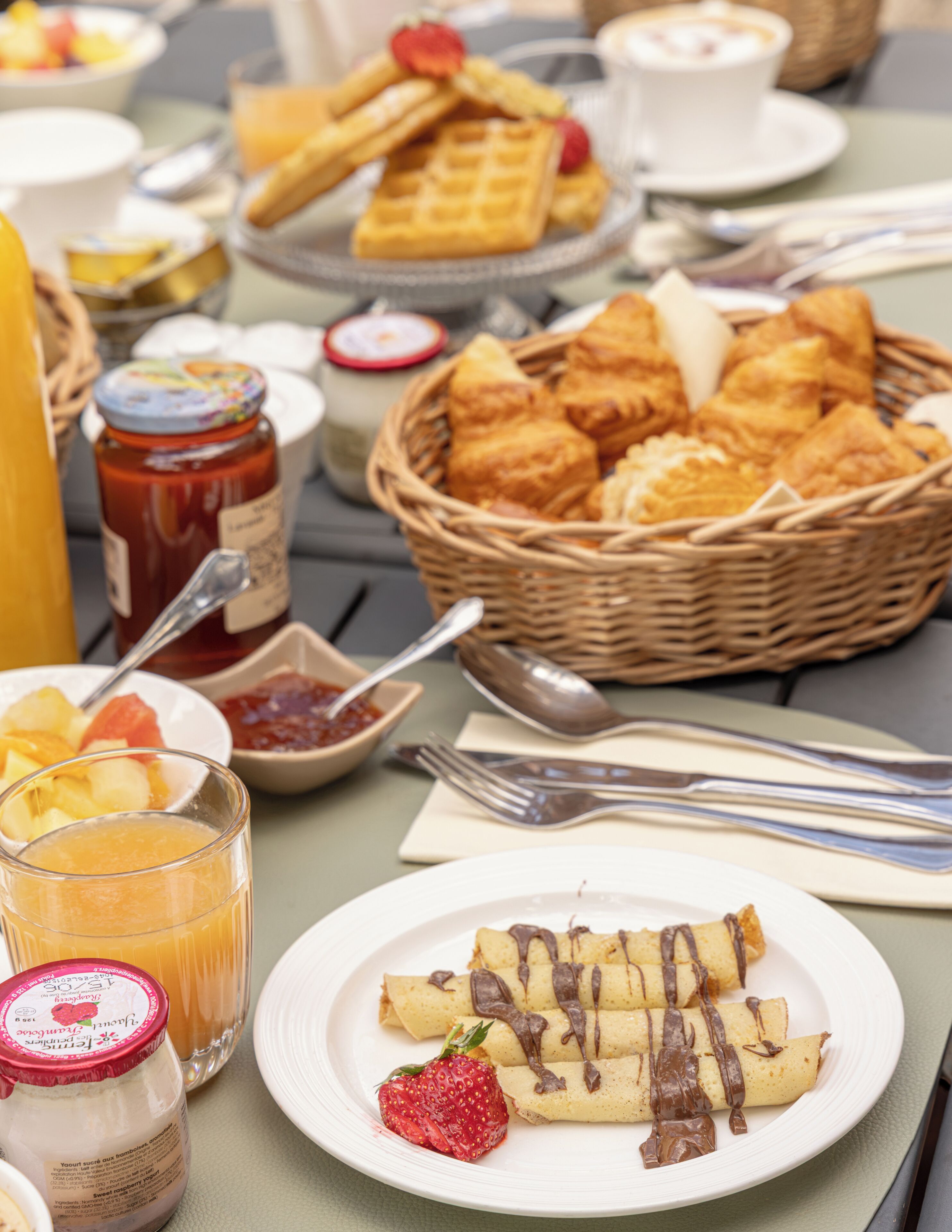 Uma mesa de café da manhã com diversos alimentos, incluindo waffles, doces e um prato de crepes com calda de chocolate.