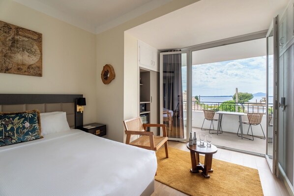Habitación de hotel con cama, escritorio, silla, una mesita con vasos y balcón con vista al mar.