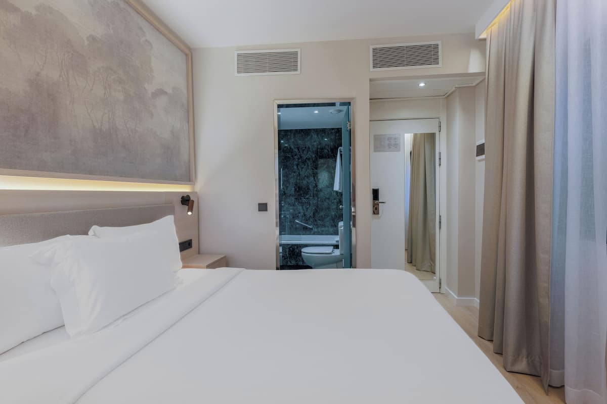 Habitación de hotel con una cama grande, un cabecero, un cuadro en la pared, un baño visible a través de una puerta de cristal y una puerta que conduce a otra habitación.
