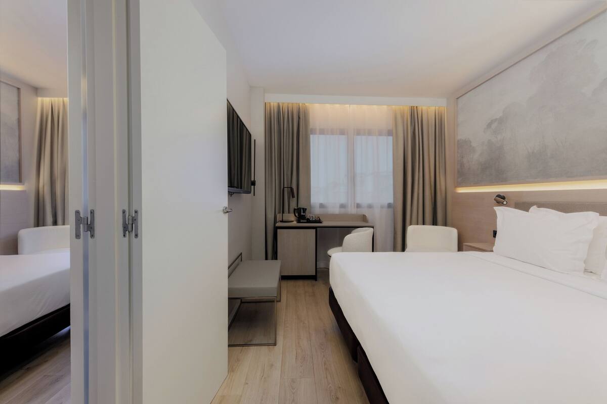 Una habitación de hotel con cama, escritorio, silla, televisor y ventana con cortinas.