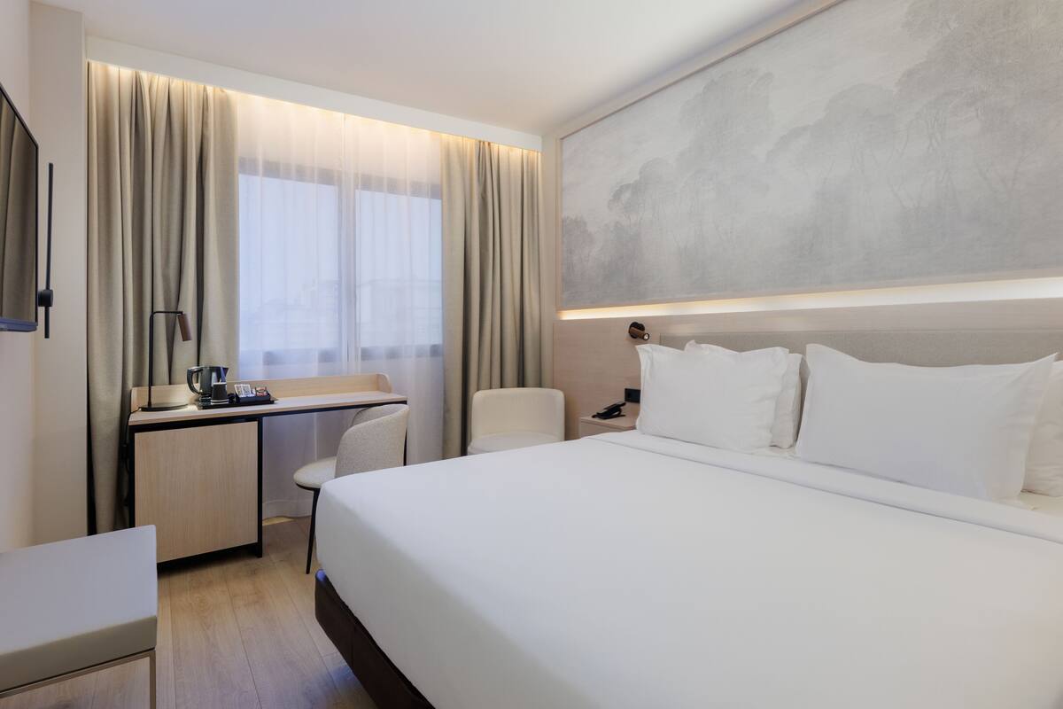 Una habitación de hotel moderna con una cama grande, un escritorio con silla, una cafetera y una mesita de noche con lámpara.