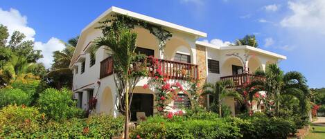 A two-story building with a balcony, surrounded by lush greenery and flowering plants.