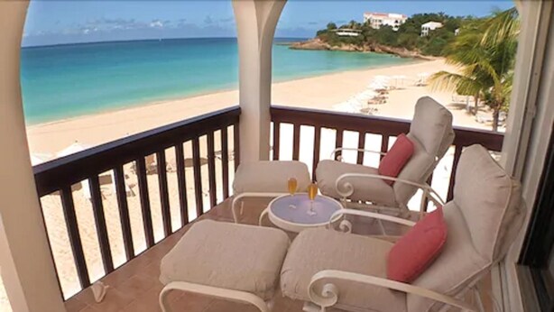 A balcony with two cushioned chairs and a small table, overlooking a beach with clear turquoise water and a sandy shore. - Carimar Beach Club (West End Village)