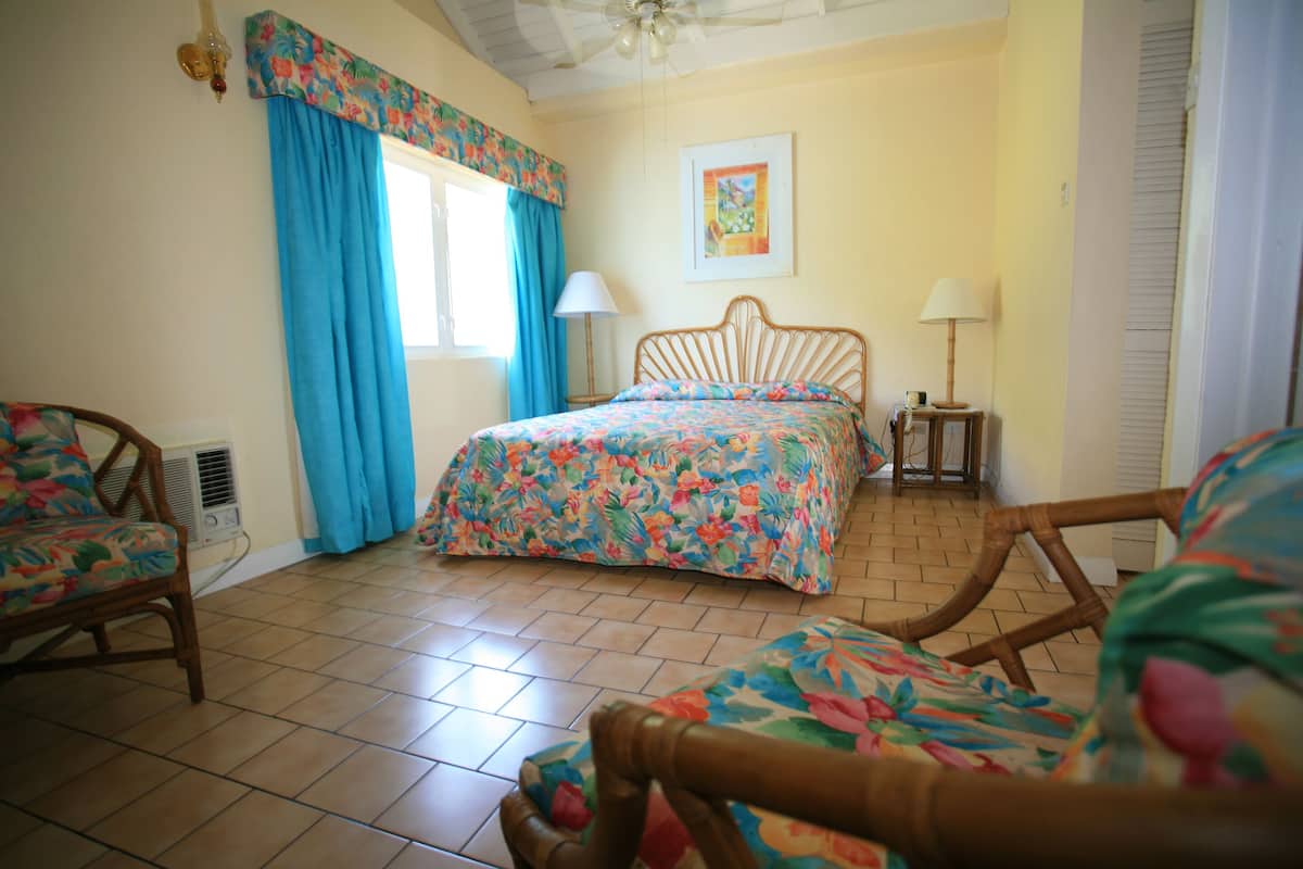 A bedroom with a bed, two chairs, a window with curtains, a bedside table with a lamp, and a wall-mounted picture.