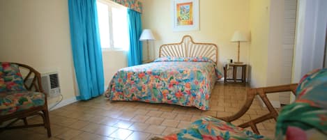 A bedroom with a bed, two chairs, a window with curtains, a bedside table with a lamp, and a wall-mounted picture.