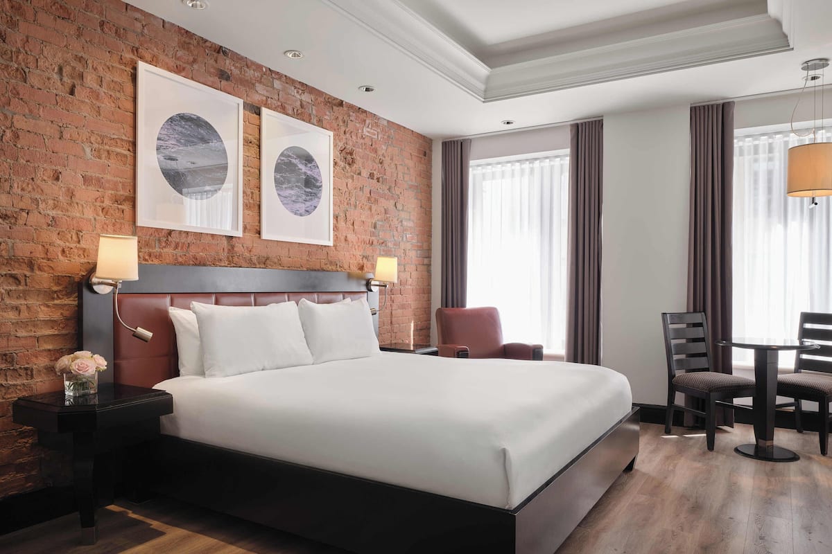 A hotel room with a brick wall, a large bed, two side tables with lamps, a desk with chairs, and two framed pictures on the wall.