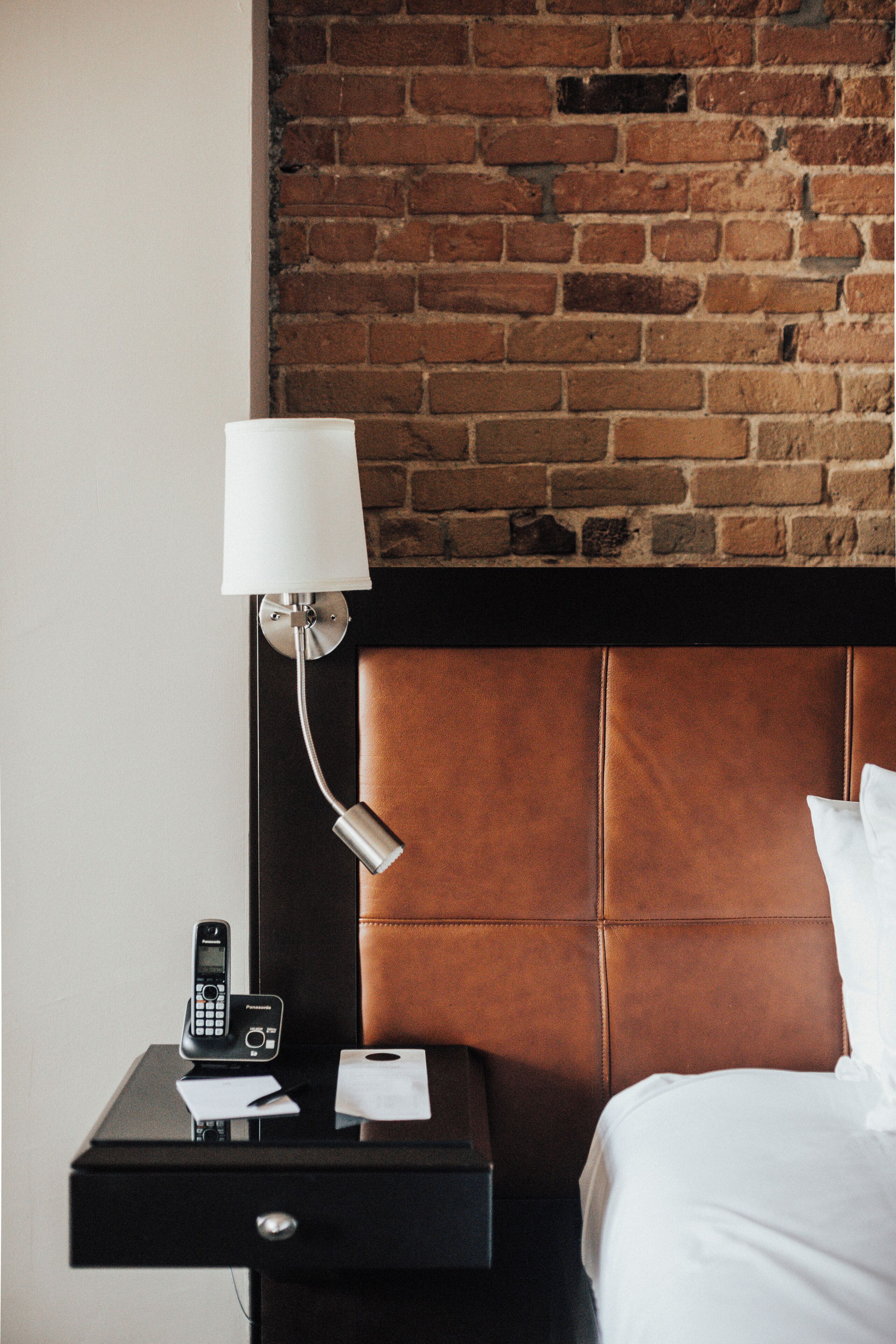 Habitación de hotel con pared de ladrillo, cabecera marrón, mesita de noche con lámpara, teléfono y bloc de notas.