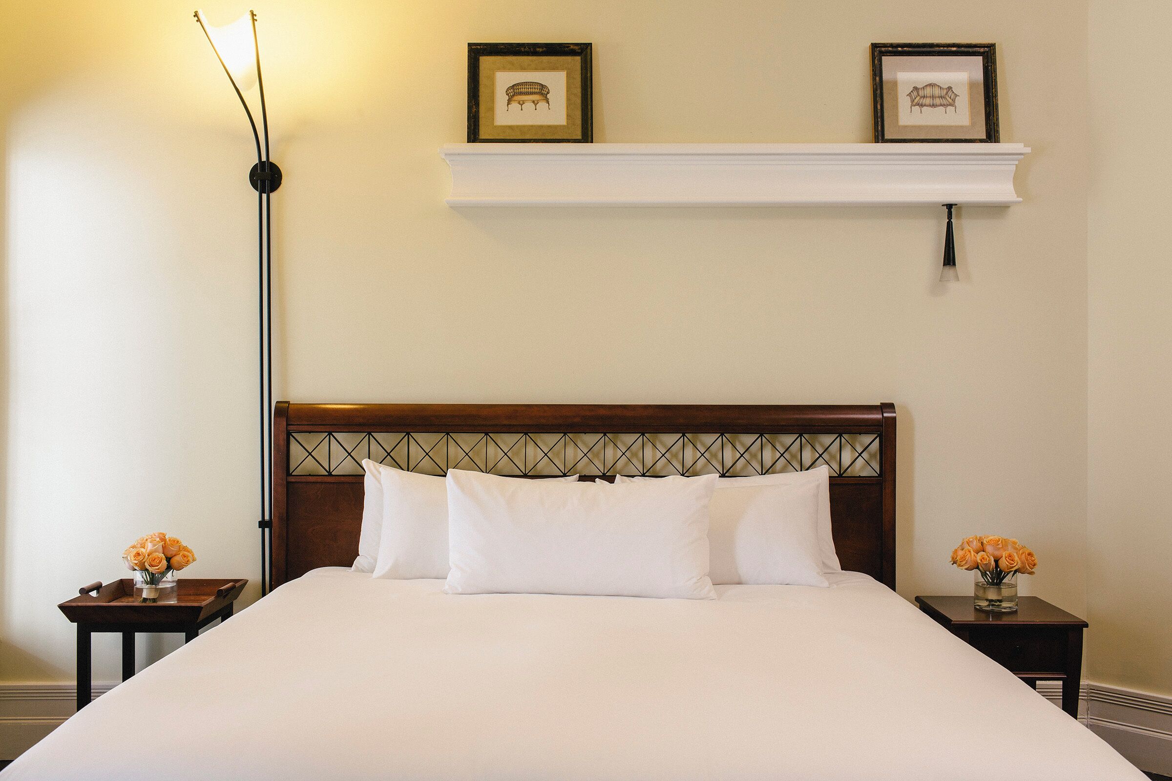 A neatly made bed with white linens, a wooden headboard with a diamond pattern, and a white nightstand with a vase of flowers.