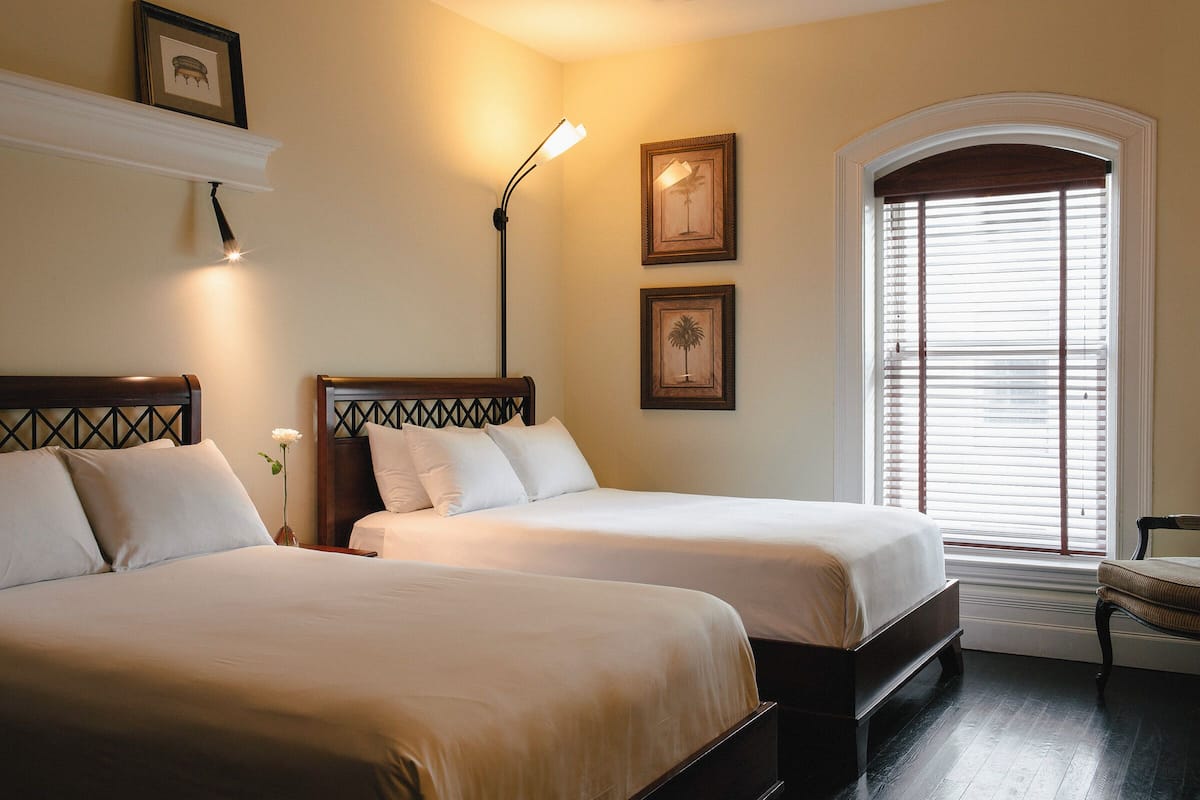 A hotel room with two beds, a nightstand, a lamp, framed pictures on the wall, and a window with blinds.
