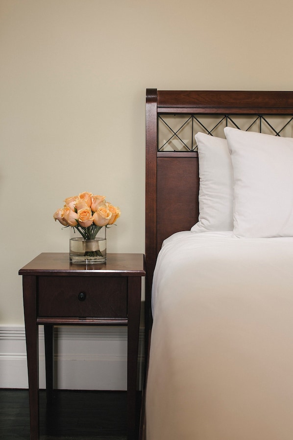 Una cama bien hecha con ropa blanca y un cabecero de madera. Una mesita de noche de madera con un cajón y un florero están colocados junto a la cama.