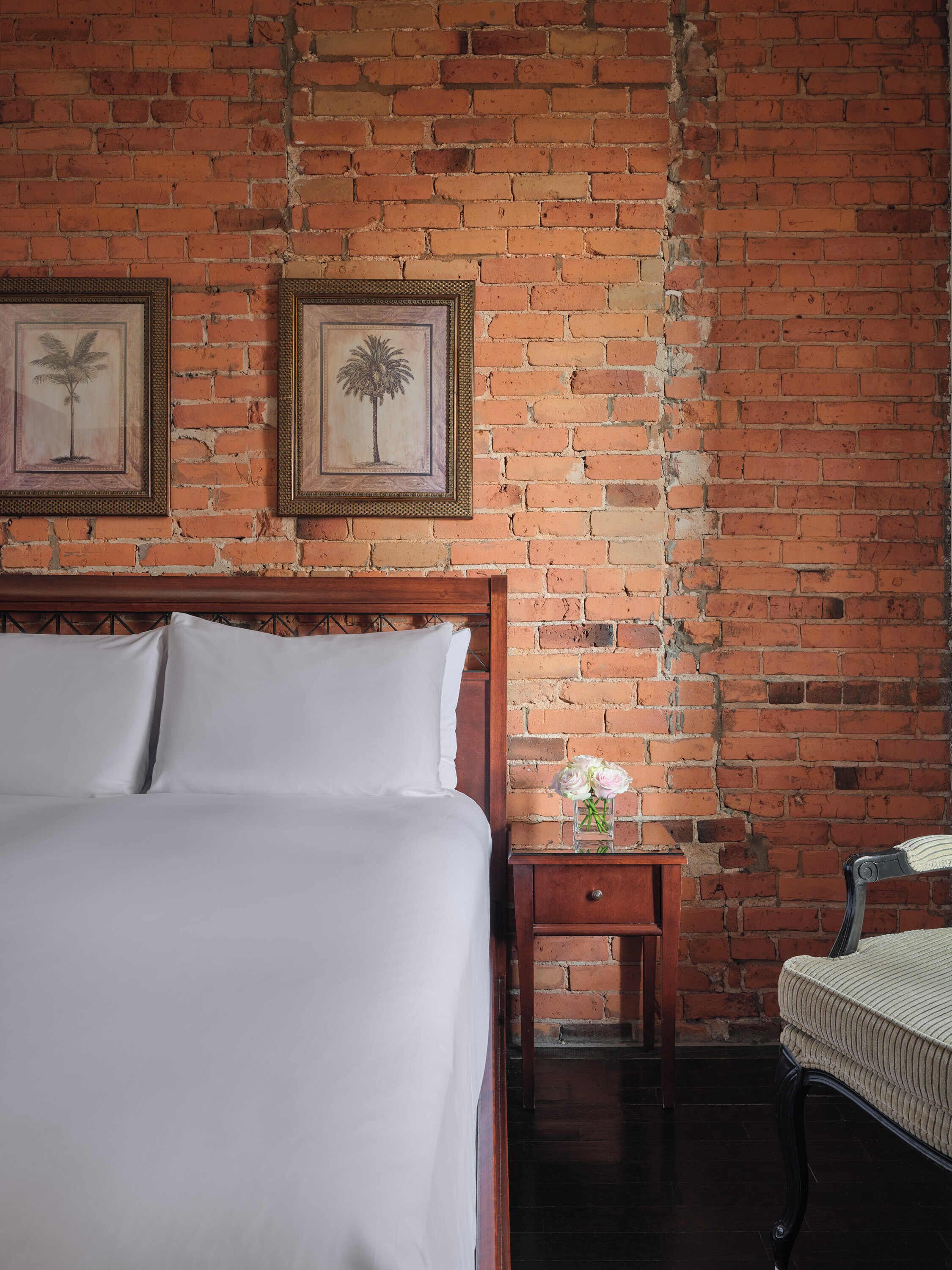 Una habitación con pared de ladrillo, una cama de madera con ropa blanca, dos cuadros enmarcados, una mesita de noche de madera con un jarrón de flores, y una silla.