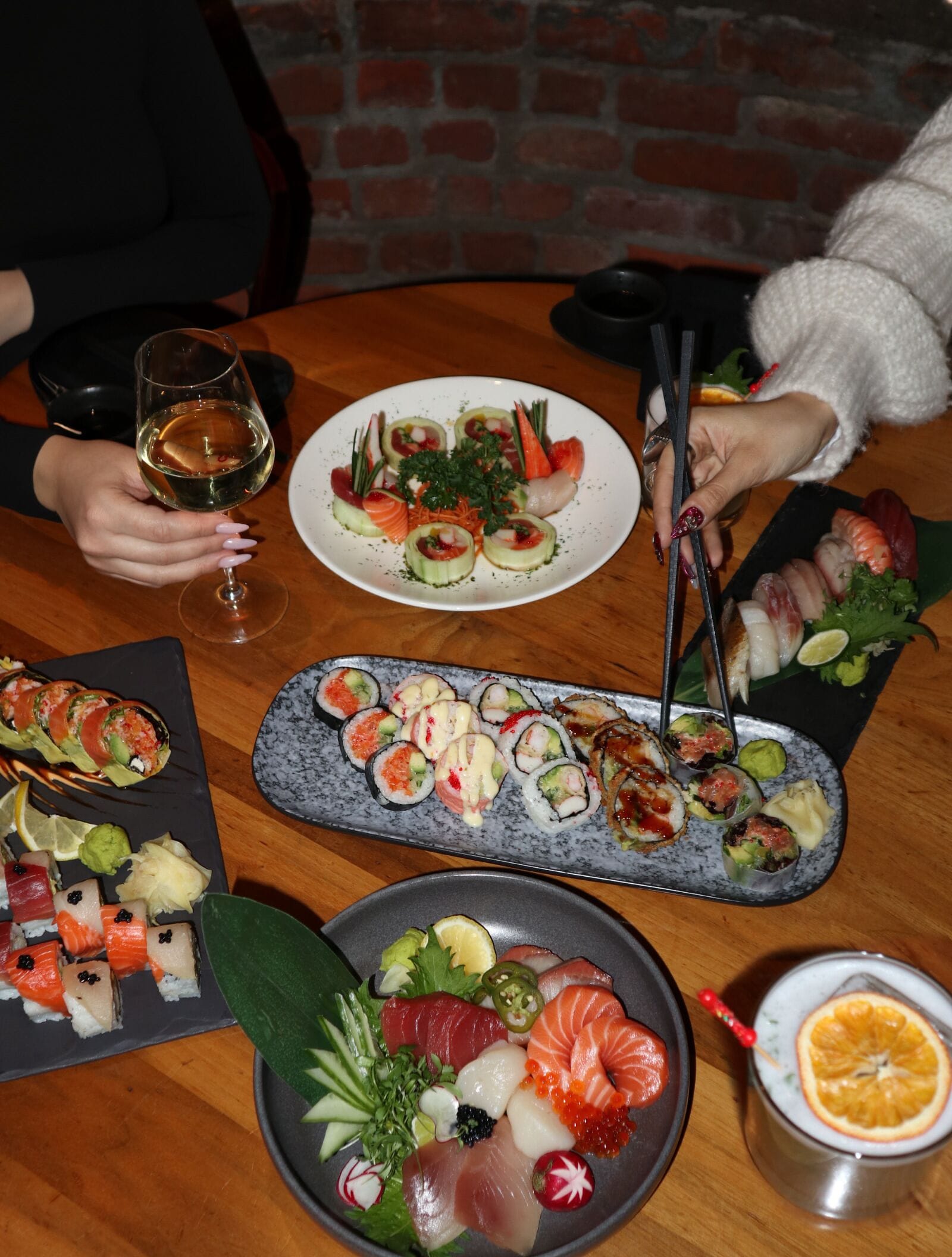 A table with various sushi dishes, a glass of white wine, and a metal cup with an orange slice.
