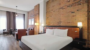 A hotel room with a large bed, a brick accent wall, a desk with a chair, and a window with curtains. - Hotel Place d'Armes (Montreal)