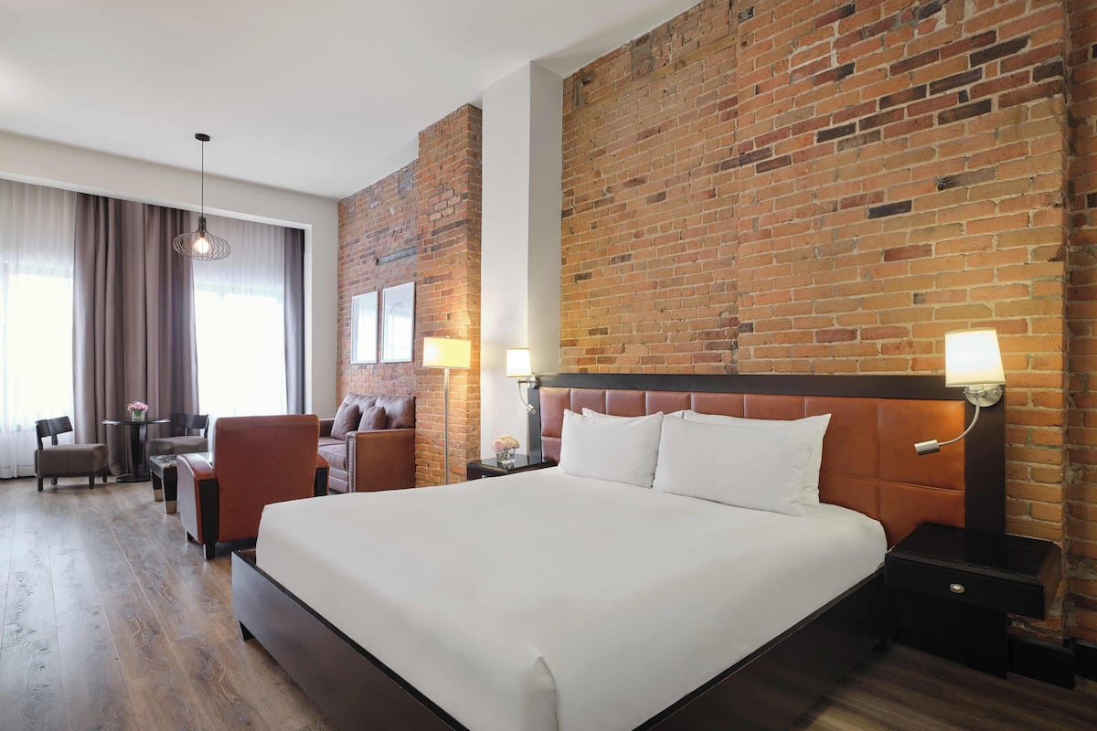 A hotel room with a large bed, a brick accent wall, a desk with a chair, and a window with curtains.