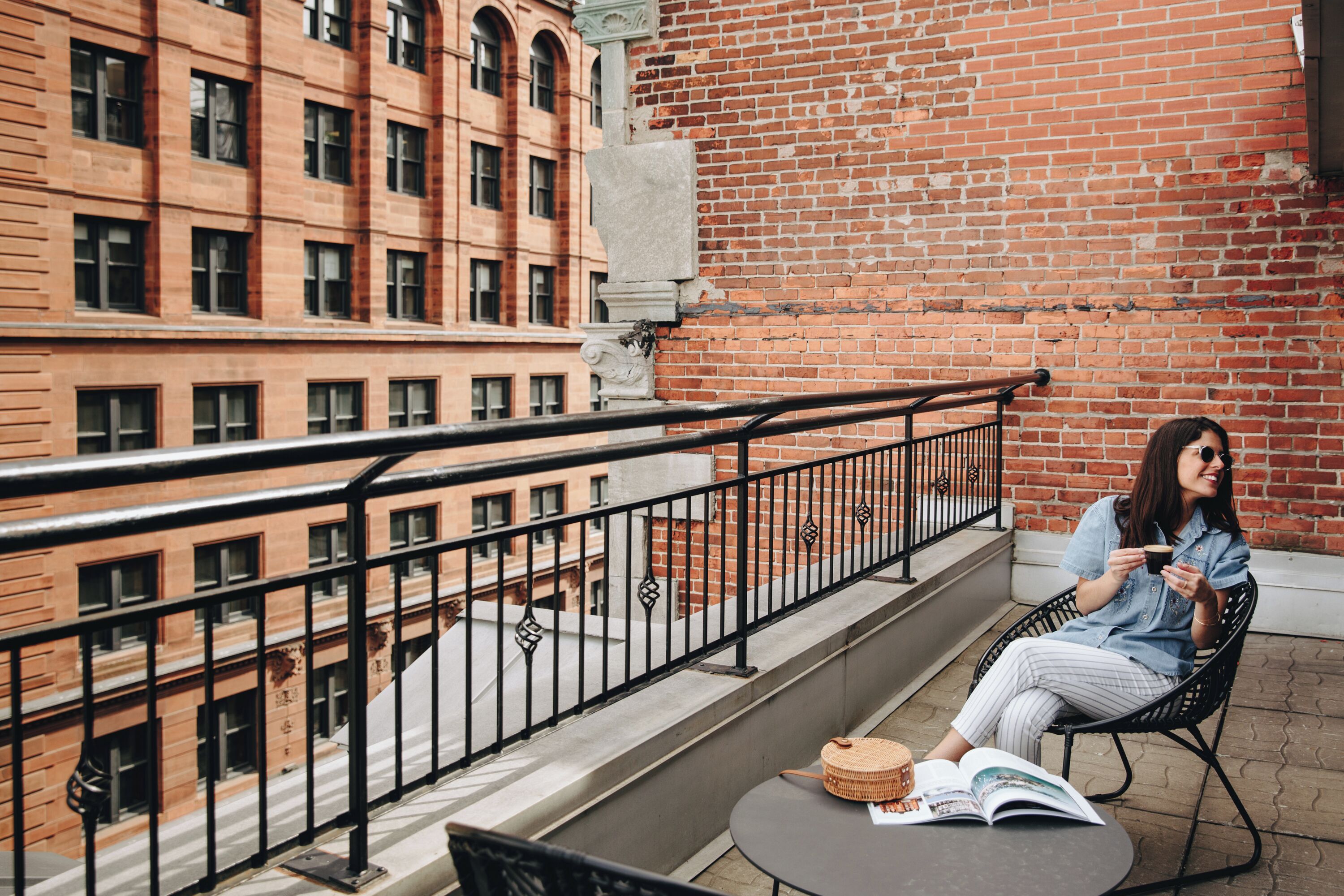Un balcón con vistas a un edificio de ladrillo, una mesa con un libro y una cesta, y una persona sentada en una silla.